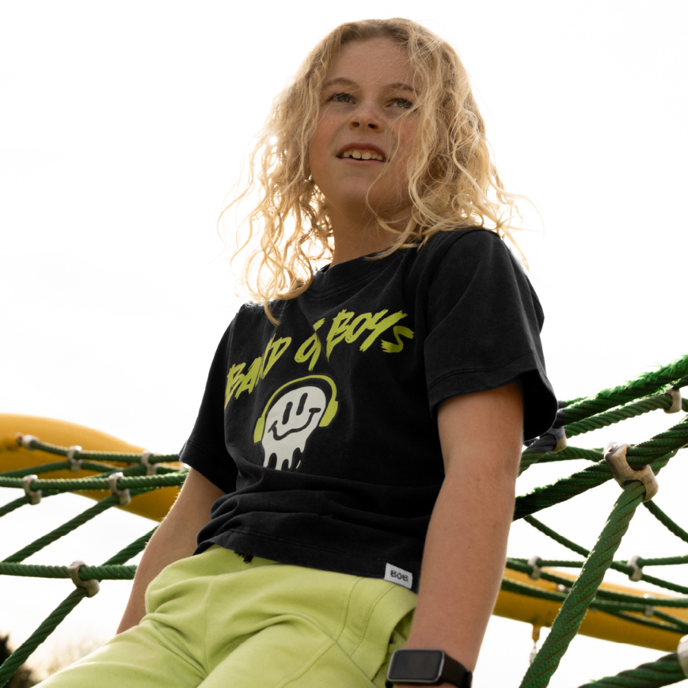 Child wearing a black t-shirt with yellow text and green shorts sitting on playground equipment.