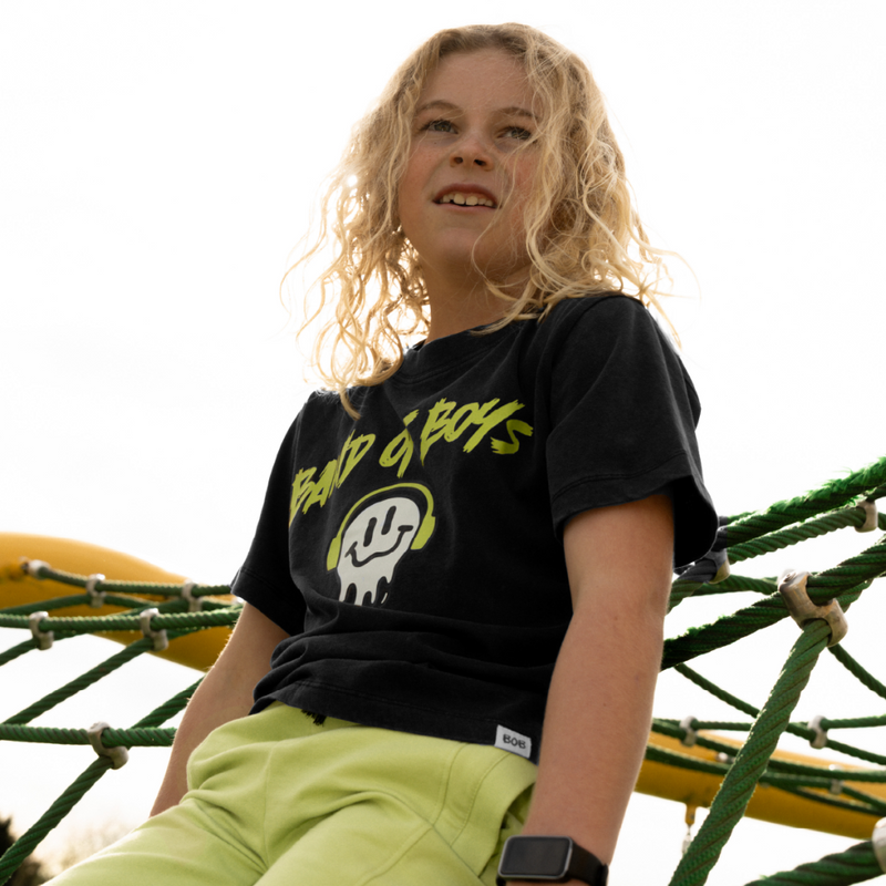 Child wearing a black t-shirt with yellow text and green shorts sitting on playground equipment.