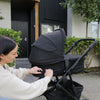 Woman adjusting a stroller in front of a modern house with greenery.
