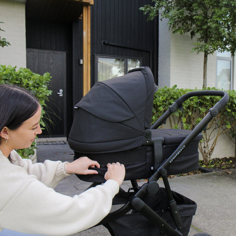 Woman adjusting a stroller in front of a modern house with greenery.