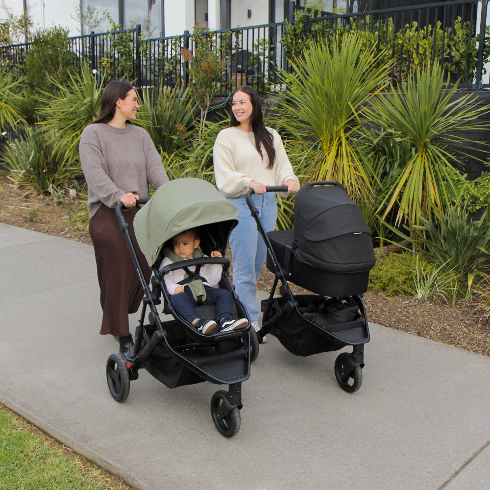 Two women pushing strollers on a sidewalk with a modern building in the background