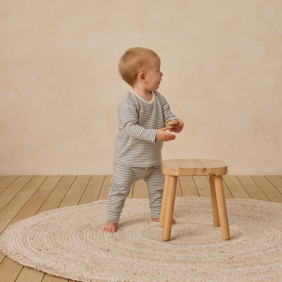 Child in a striped outfit standing on a wooden floor with a round rug and small wooden stool.
