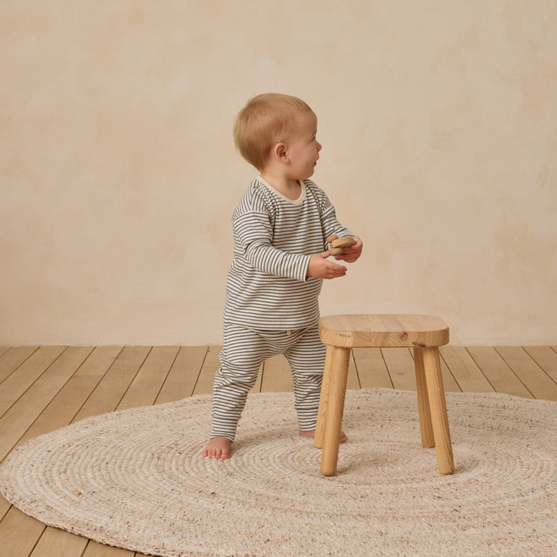Child in a striped outfit standing on a wooden floor with a round rug and small wooden stool.