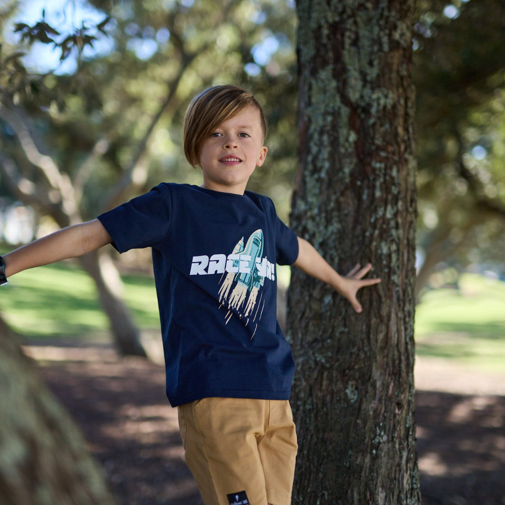 Child wearing a blue t-shirt with a graphic design, standing next to a tree in a park.