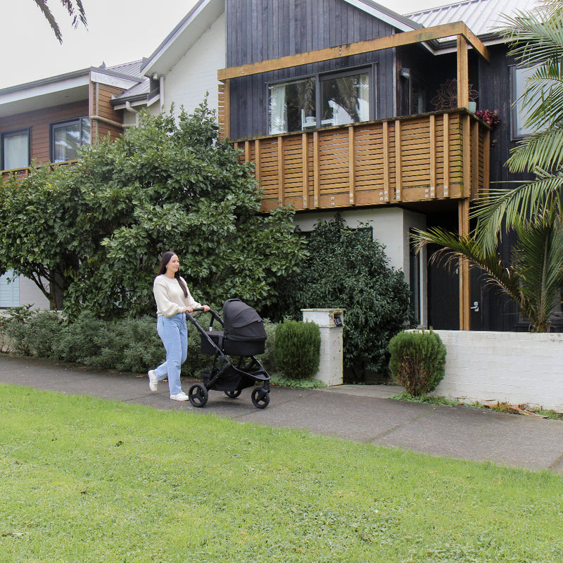 Woman pushing a stroller on a sidewalk in front of a modern house with greenery.
