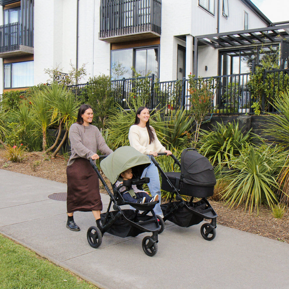 Two women pushing strollers with children on a sidewalk in an urban setting.