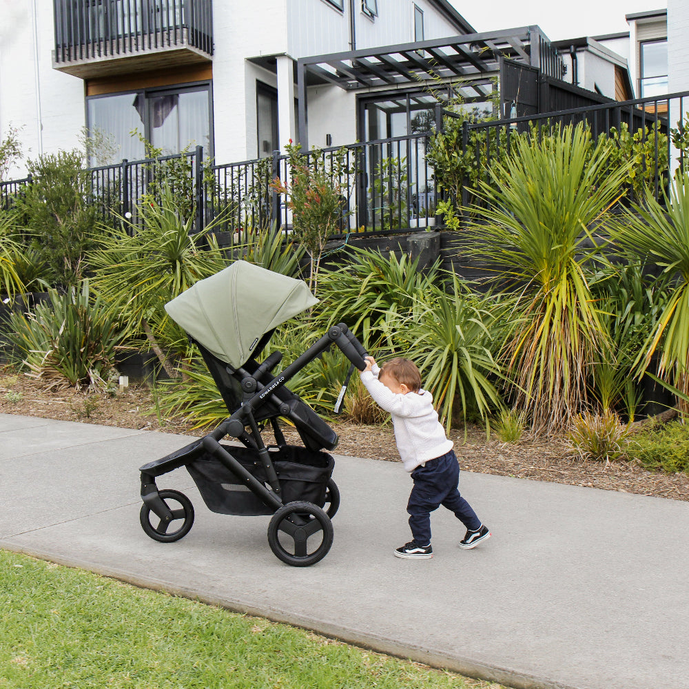 Child pushing a stroller on a sidewalk with greenery and buildings in the background