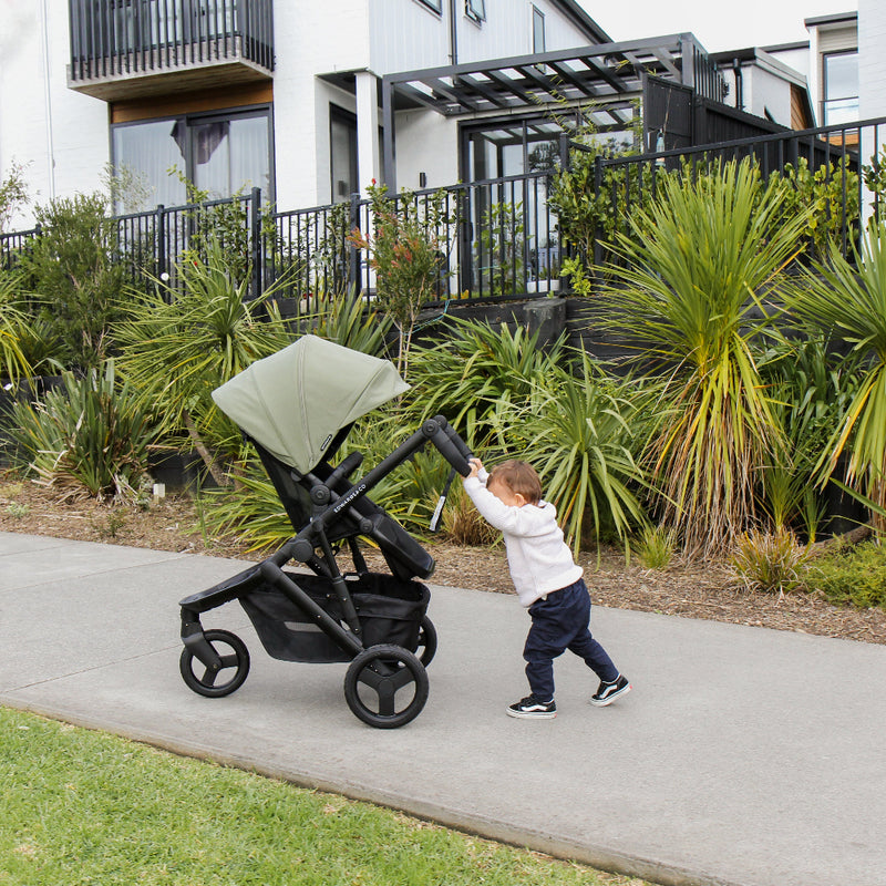 Child pushing a stroller on a sidewalk with greenery and buildings in the background